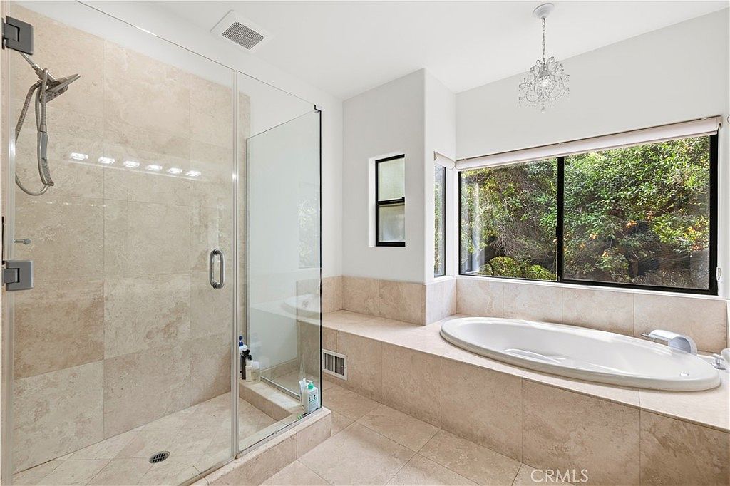 Primary bath with travertine surround climbing the wall, deep soaking tub framed by glass, and a small crystal pendant overhead
